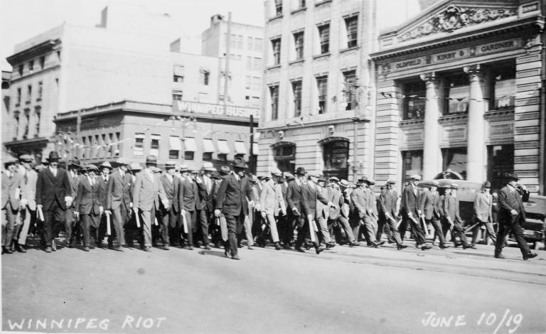 A large group of men in suits and hats brandishing clubs marching down the street.
