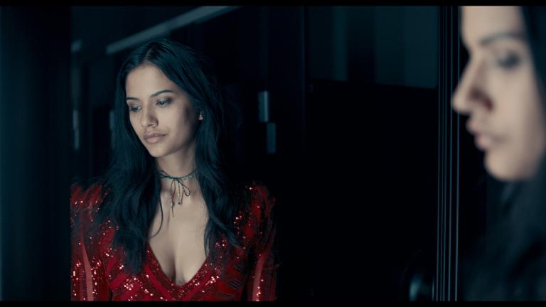 A woman with long dark hair and a red dress is pictured from the waist-up, looking downward towards something off screen. Another woman's face can be seen on the other side of the frame, looking in the same direction. Partially obscured.