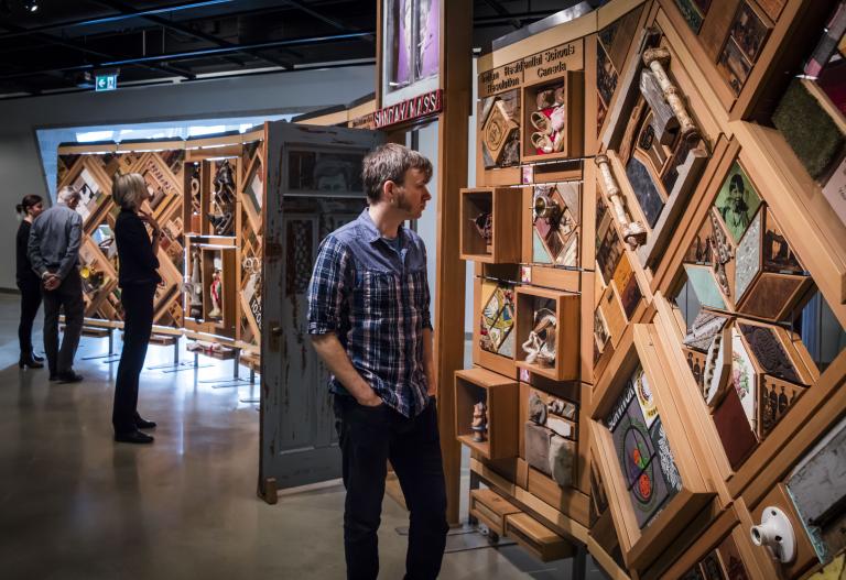 Two people look at a large artwork consisting of objects set in cedar frames, with an open door in the middle. Partially obscured.