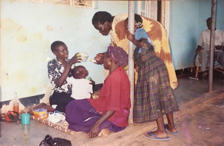 Two women are sitting on the ground together near a wall inside a building. One is holding a baby and taking a white object from a third woman. A young girl stands next to the third woman.