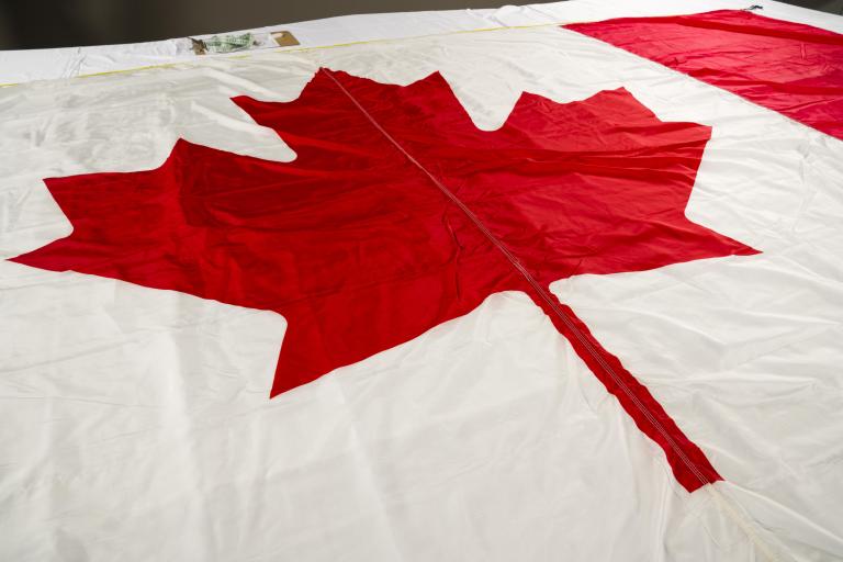 A close-up image of a Canadian flag, with a focus on the red maple leaf. Partially obscured.