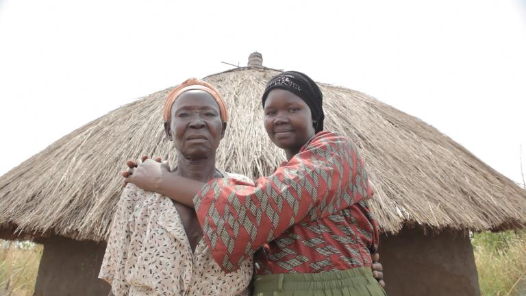 Two women are standing in front of a hut with a straw roof, looking at the camera. The younger woman on the right is smiling and embracing the one on the left. Partially obscured.