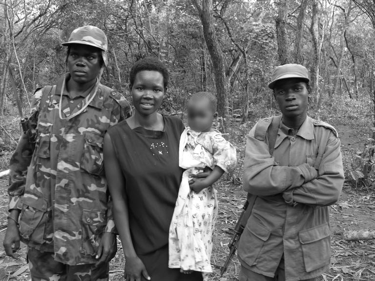A black and white photograph of a woman holding a baby, while two men in army fatigues stand beside her. They are standing in front of a forest, posed for the camera.