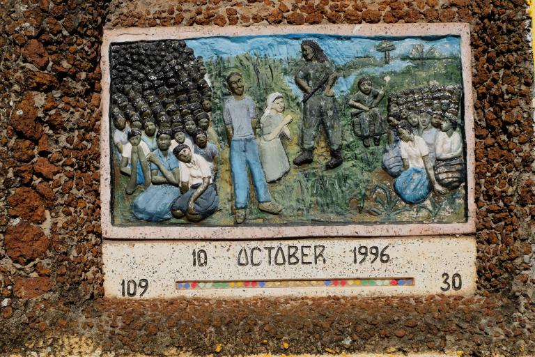 An art piece that appears to be made out of stone and clay. A man with a gun stands in the middle, with girls on their knees all around him. Another man stands with his hands behind his back, as if handcuffed, and a woman wearing a white outfit kneels in front of the man with a gun, praying.