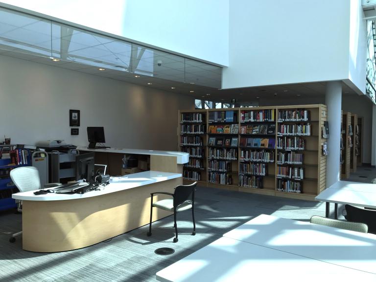 A room containing books on bookshelves, a reference desk with a computer workstation and worktables. Partially obscured.