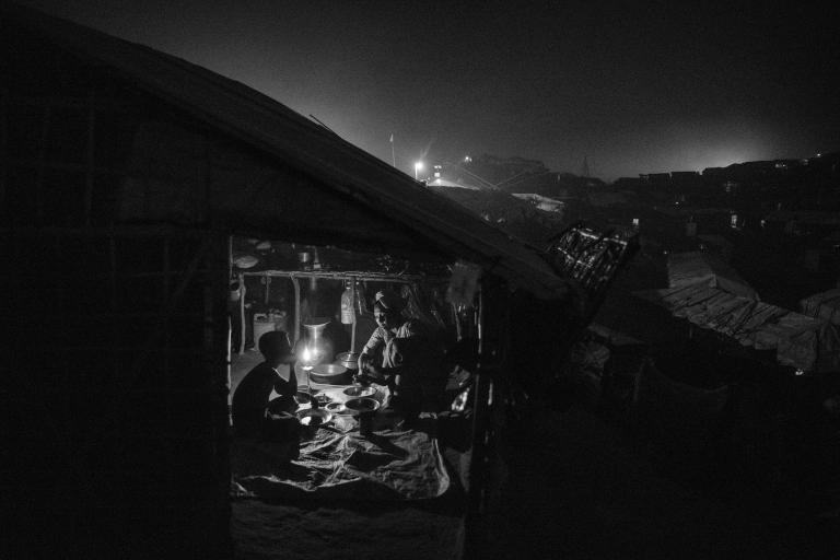A Rohingya refugee woman serves food to a boy who sits across from her in a dark shelter. The interior is lit by a single candle.