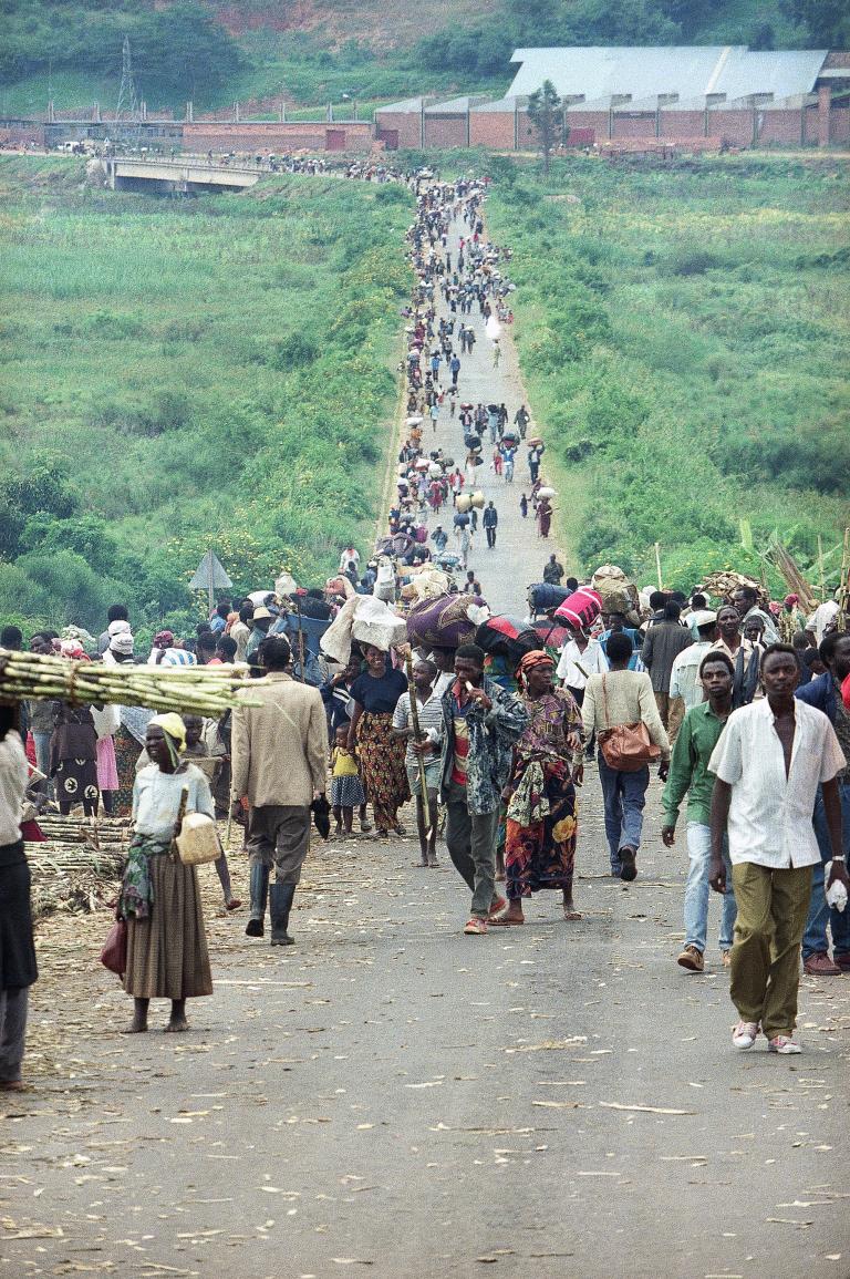 A large crowd of people of all ages carrying food and belongings walk toward the camera on a long dirt road through a bright green landscape of grass and bushes. The road and the crowd extend far into the distance. Partially obscured.