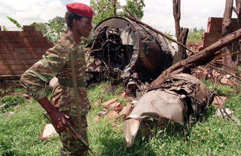 A soldier carrying a rifle and wearing a red beret and camouflage uniform stands in front of the wreckage of a plane among trees.