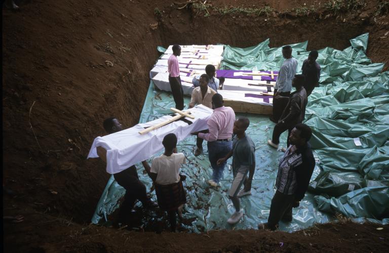 Several men carry a coffin wrapped with cloth and adorned with a large wooden cross into a large grave dug deep into the earth that already contains several similar coffins.