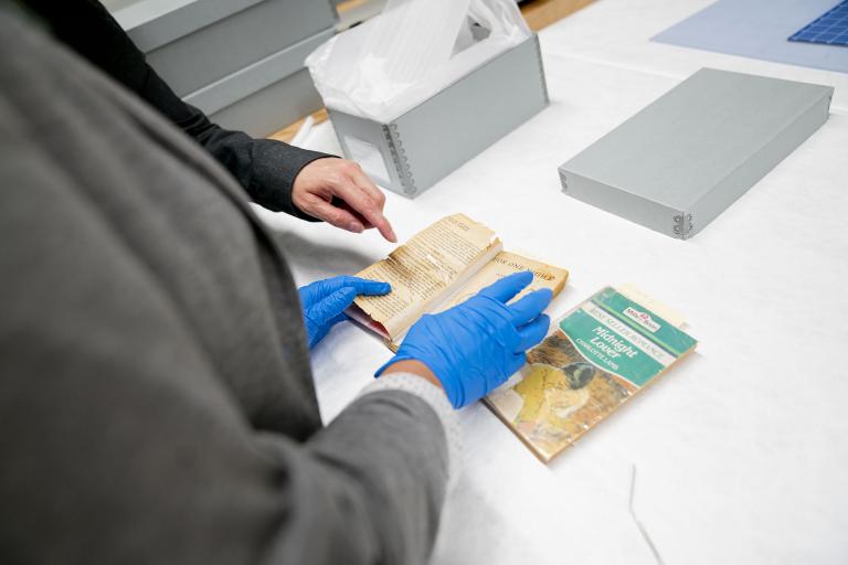 Hands with blue protective gloves are holding a paperback book open to a page on a table. Another person’s hand is pointing to something in the open book. Another book also sits on the table, as well as a metal box and lid.