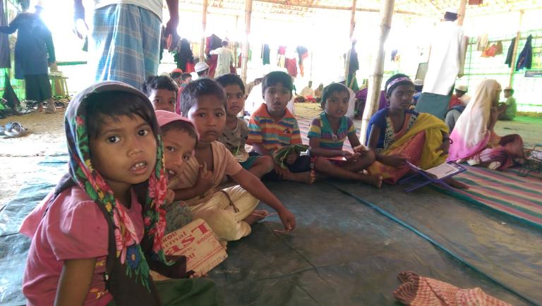 A group of children wearing colourful clothes sit cross-legged on the ground inside a shelter. Adults stand behind them, and there are clothes hanging on walls.