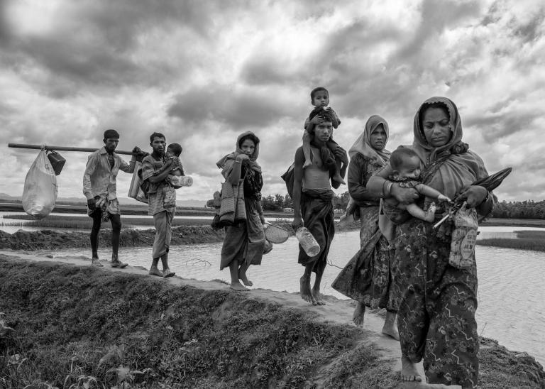 A group of Rohingya women and men carry young children and belongings as they walk in a line over an earthen dike over a stretch of water. Partially obscured.