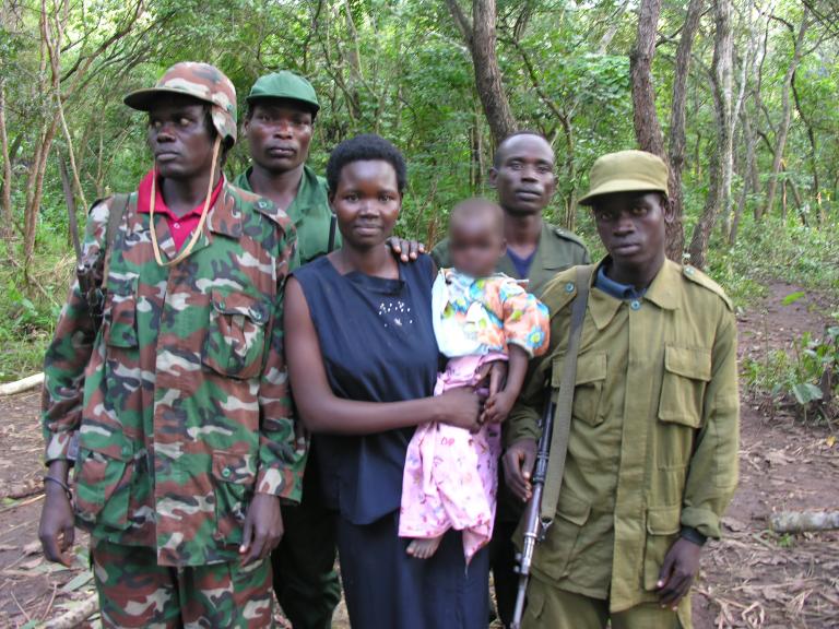 A woman is holding a baby, while four men in army fatigues stand beside and behind her. They are all standing in front of a forest, posed for the camera. Partially obscured.
