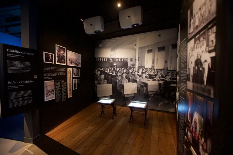 A Museum exhibit showing a black-and-white photo of children sitting in rows at school desks. Two desks, similar to those in the photo sit in the centre of the exhibit. A headline on a text panel reads “Childhood Denied.” Partially obscured.