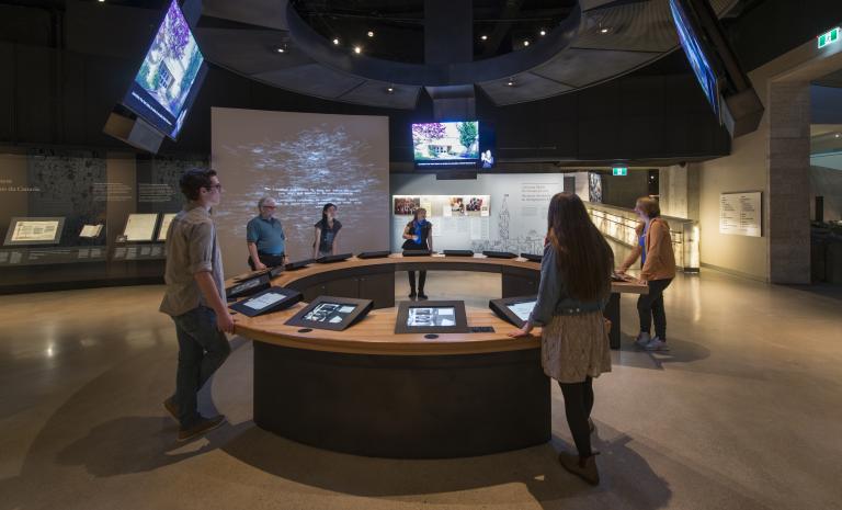 Six people in a museum gallery stand around a circular counter with video screens both embedded in the counter and hanging above them. Partially obscured.