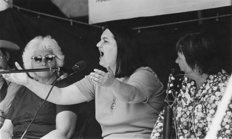 A black and white photo of a woman singing into a microphone with her arms raised. Another woman sits beside her and watches.