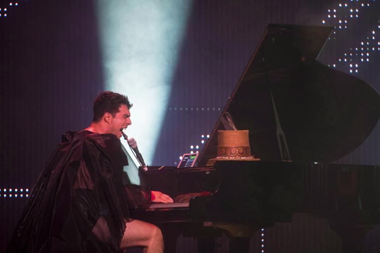 A man wearing a sheer black cape and black shorts is playing a grand piano and singing intensely into a microphone. A spotlight shines towards the ceiling behind him.