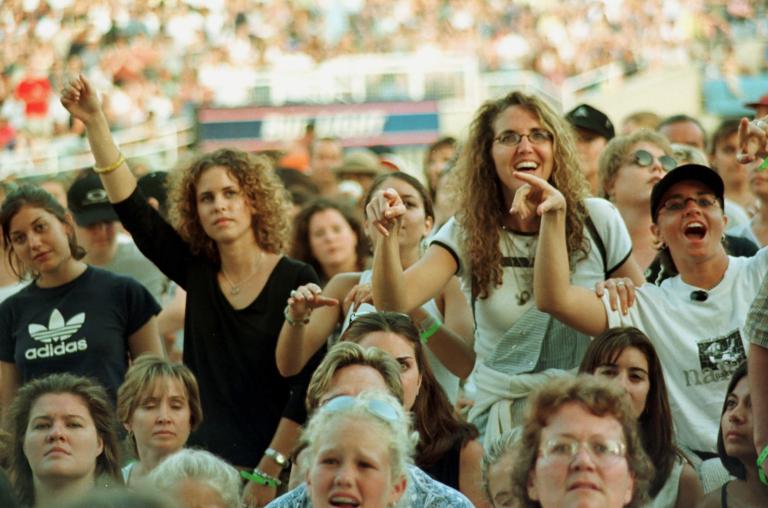 A crowd of women, viewing some sort of entertainment off-camera. Many are smiling and some have their arms raised. A larger crowd can be seen seated in a stadium behind them.