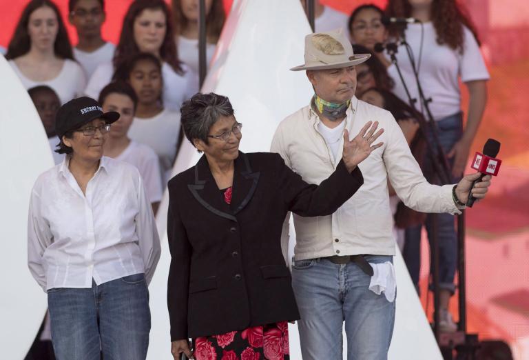 Two women and a man stand on a stage while a group of people in white shirts stand on risers behind them. The woman in the centre is waving to someone off-camera, and the man on the right is holding a microphone. Partially obscured.