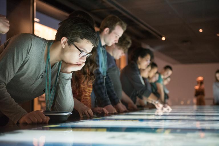 A young person leaning on a digital table reading exhibit content. In the background a Museum staff person shows a group how to use the table. Partially obscured.