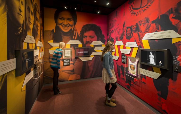 Two people reading text on a ribbon that runs along all three walls of an exhibit alcove, which also features three video screens and small artifact cases. Large historical photos with yellow, orange and red tint show women’s faces, a smiling woman and man, and women protesting. Partially obscured.