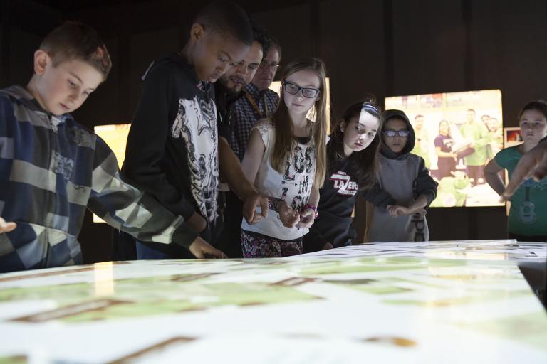 A Museum guide showing five students how to engage with the digital table, around which they are all standing. Partially obscured.