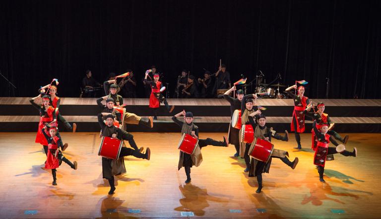 A drumming troupe dancing on a stage. The drums and parts of their costumes are a bright red. Partially obscured.