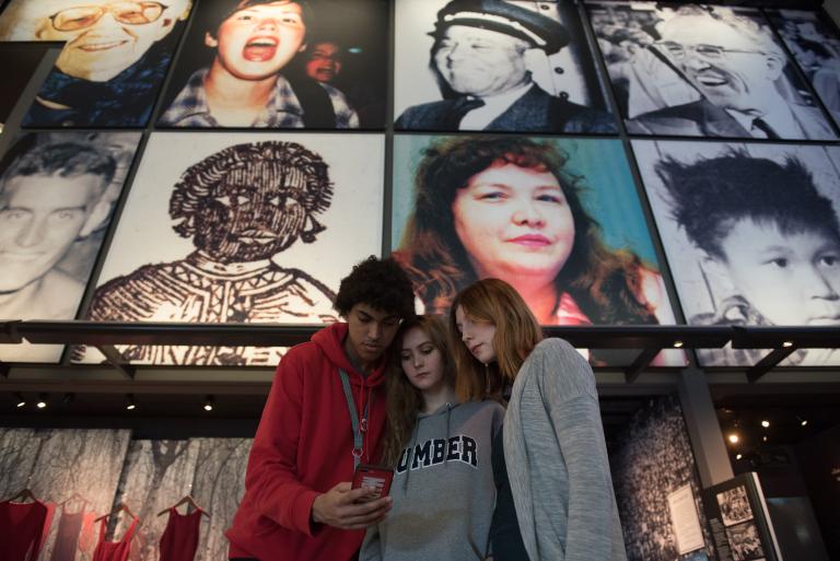 Three youth looking at a phone together. Behind them are Museum exhibits including hanging red dresses and a grid of large photos of people’s faces. Partially obscured.