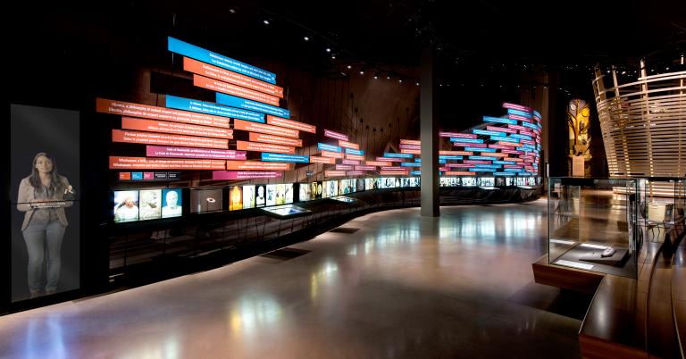 View of Museum gallery. Text on colourful rectangles on a large exhibit wall which runs the length of the gallery. Below this timeline of events is a line of backlighted photos. On the right side of the frame there is a tall exhibit element featuring a nature scene and a circular wood structure. Partially obscured.