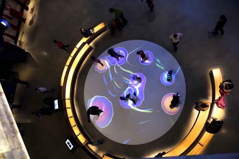 A photo looking down into a gallery. A group of about 9 people stand inside of a large circle on the floor, with colourful lights creating circles around each of their feet. People are seated on benches that surround the circle. Partially obscured.