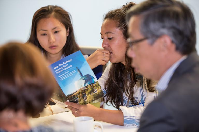 Two women are seated at a table looking at a program that says "After the Apology." Two other people are seen in the foreground seated at the same table. Partially obscured.
