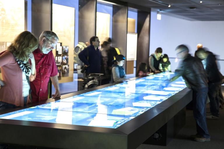 Museum visitors are looking at a large rectangular table, the surface of which is a touchscreen.