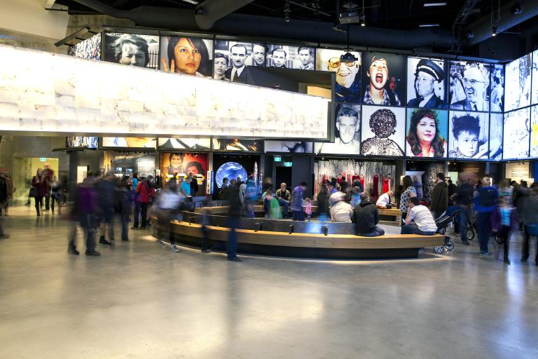 A large crowd in a museum gallery. There is a curved bench in the centre of the room. The display consists of a series of portraits above alcoves each containing a different exhibit.