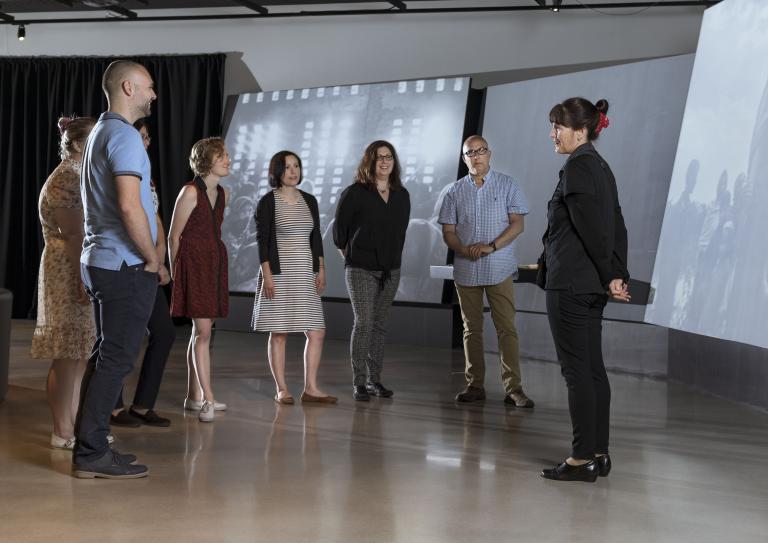 A small group of people stand in a semicircle in front of large screen displays. Partially obscured.