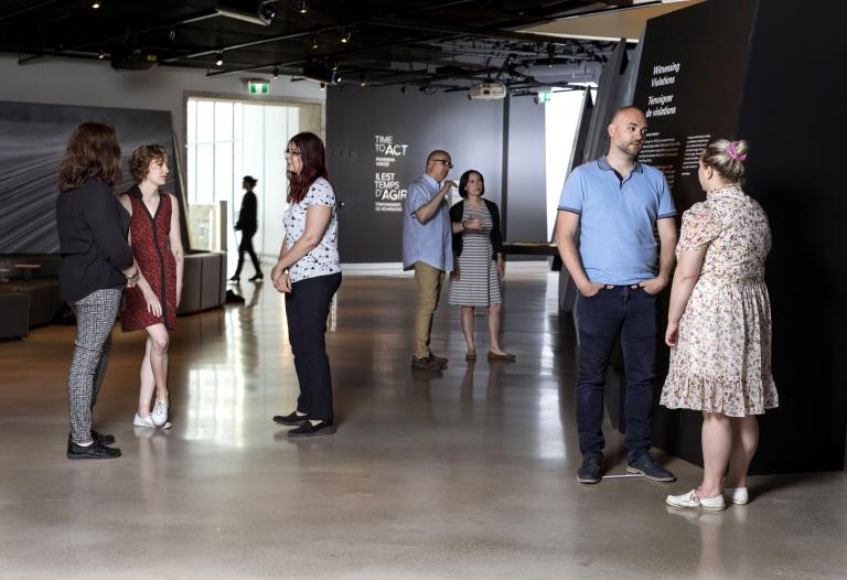 Small groups of people stand in a Museum exhibition space and talk to one another. Partially obscured.
