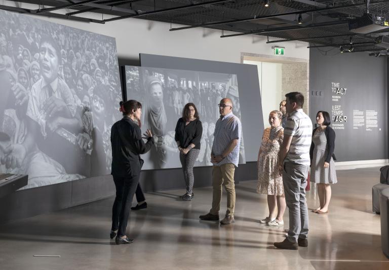 A small group of people stand in front of large screens displaying photographs and look at a woman who is speaking to them. Partially obscured.