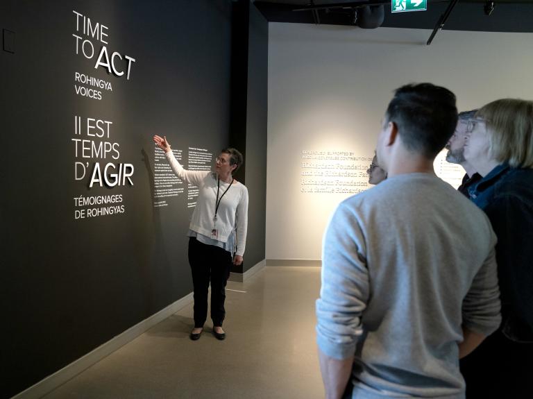 A woman points to a wall that says “Time to Act, Rohingya Voices” in both English and French while a small group of people stand and watch. Partially obscured.