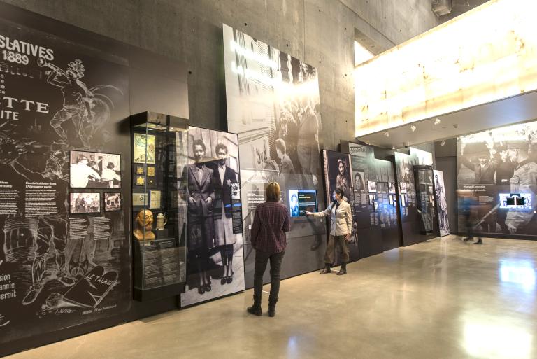 Two people explore a museum gallery containing images, artifacts, text and interactive screens.
