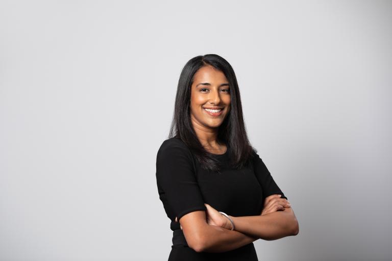 A portrait of a smiling woman standing with arms crossed in front of a white backdrop. Partially obscured.