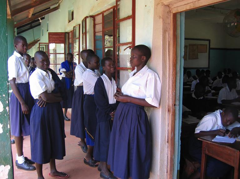 A group of young women and girls in their teens stand together on the front porch of a building. Most are dressed in white blouses and blue skirts, with two girls also wearing blue sweaters. To the right, a doorway reveals a roomful of students sitting at wooden desks and writing on notepads. Partially obscured.