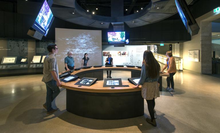 Six people in a museum gallery stand around a circular counter with video screens both embedded in the counter and hanging above them.