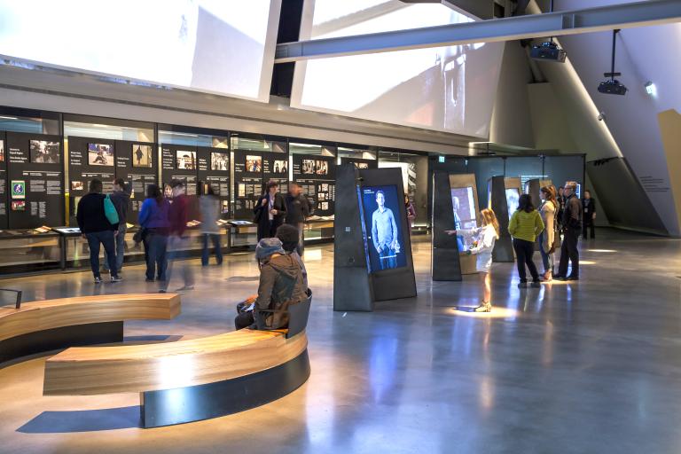 People explore a gallery containing vertical interactive videos in the centre of the room; and displays containing text and images that line the back wall.