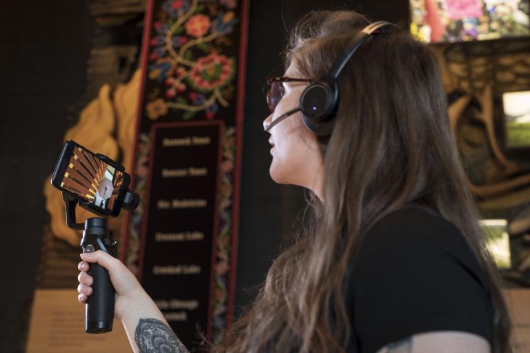 A woman holds a bar with a mobile phone attached to it, and uses the screen to look at something off camera in a Museum gallery. Partially obscured.