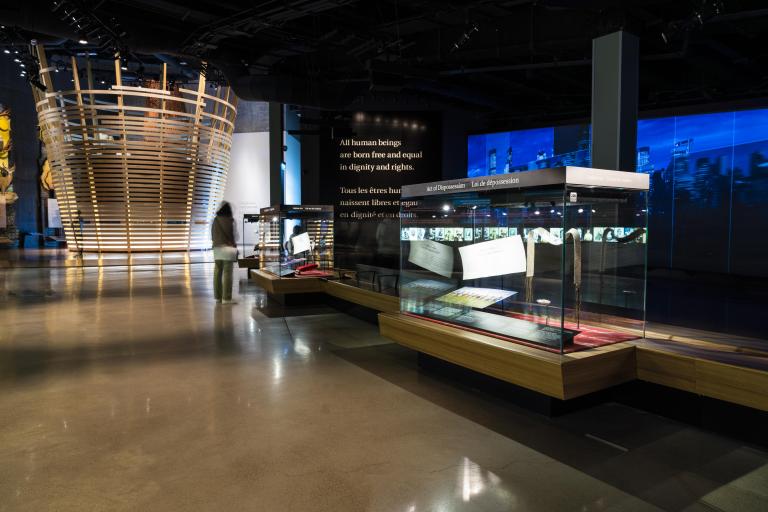 A museum gallery containing a large circular structure with horizontal wooden slats; glass cases containing artifacts; and projections on the wall. Partially obscured.