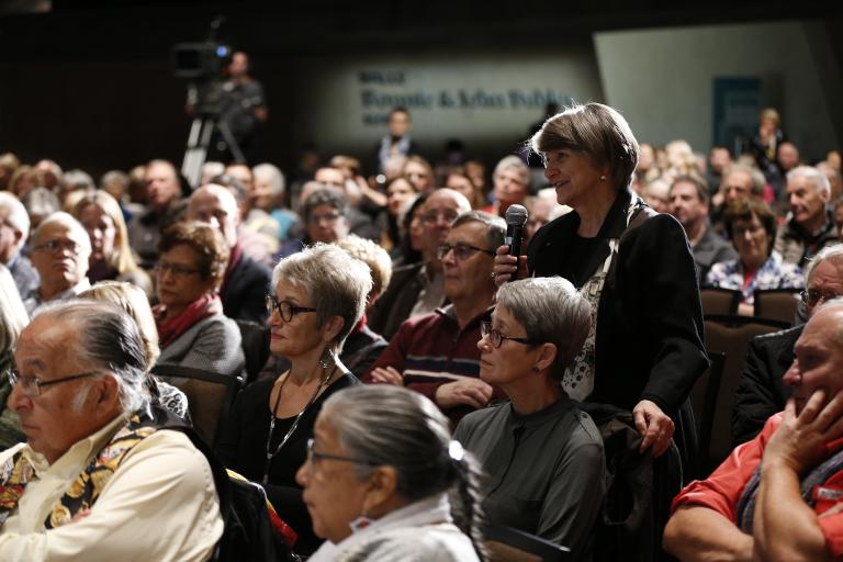 A room full of audience members sitting. One woman is standing and smiling holding a microphone. Partially obscured.