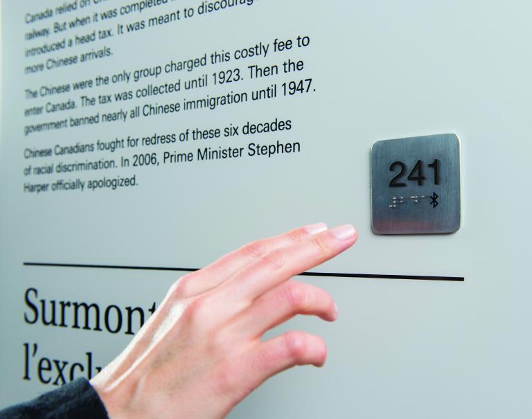 A hand reaches out to a Universal Access Point, which is a small metal square attached to a wall. The square has the number 241 imprinted on it, as well as braille writing and a Bluetooth symbol.