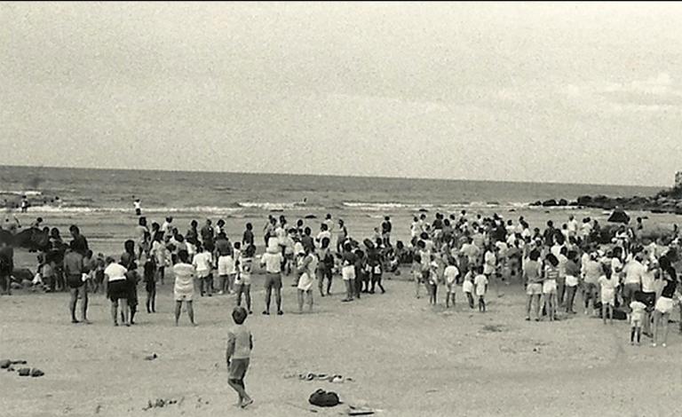 A black-and-white photograph of a crowd of people, most of them standing, on a beach. Partially obscured.