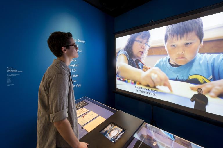 A man stands in front of a screen playing a film that shows an adult working with a child. English subtitles and a sign language interpreter are also displayed on the screen. Partially obscured.