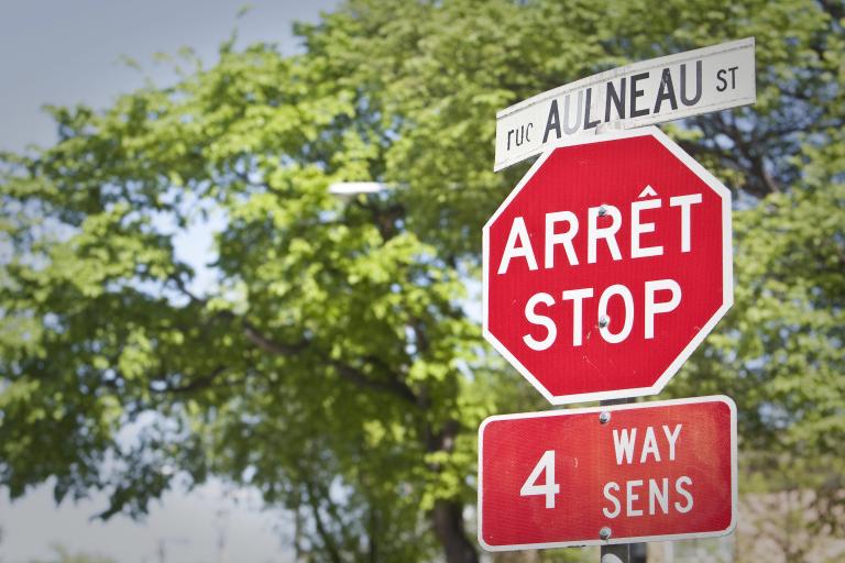 A stop sign and street sign in both English and French are seen in front of trees. Partially obscured.