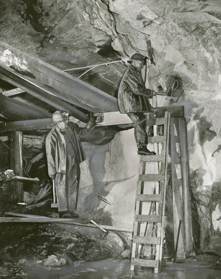 Two miners wearing helmets work underground in a uranium mine. One stands on a ladder, as the other miner watches him.